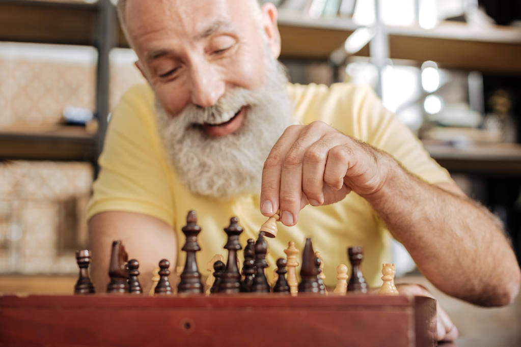 Iloinen vanhempi mies liikuttaa sotilasta shakkilaudalla
 - Valokuva, kuva