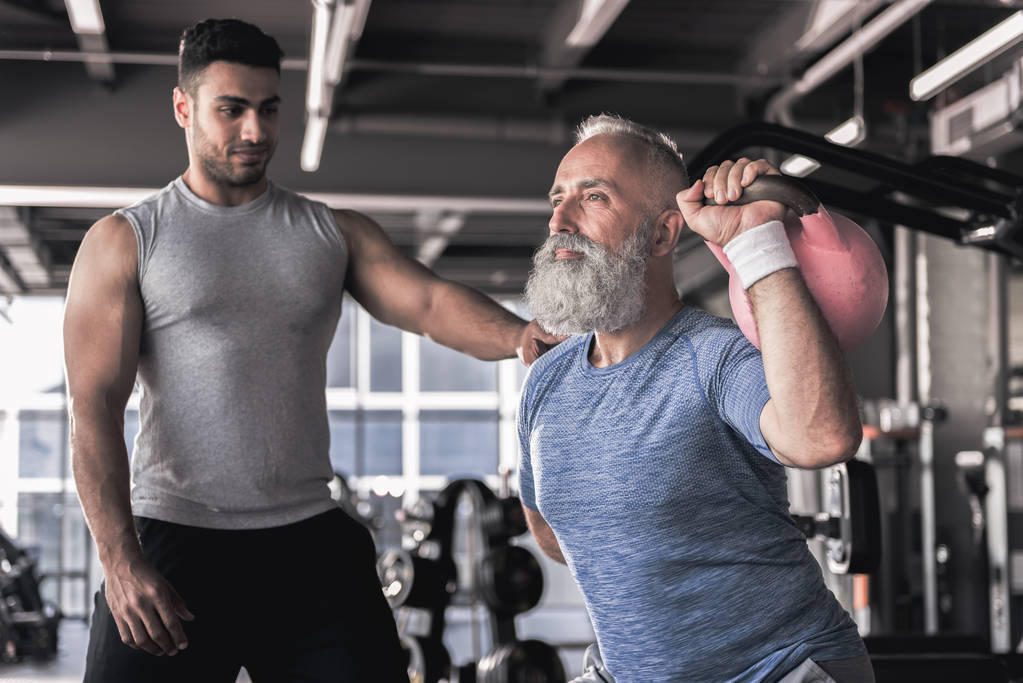 Старший мужчина делает упражнения с молодым инструктором в современном тренажерном зале
 - Фото, изображение