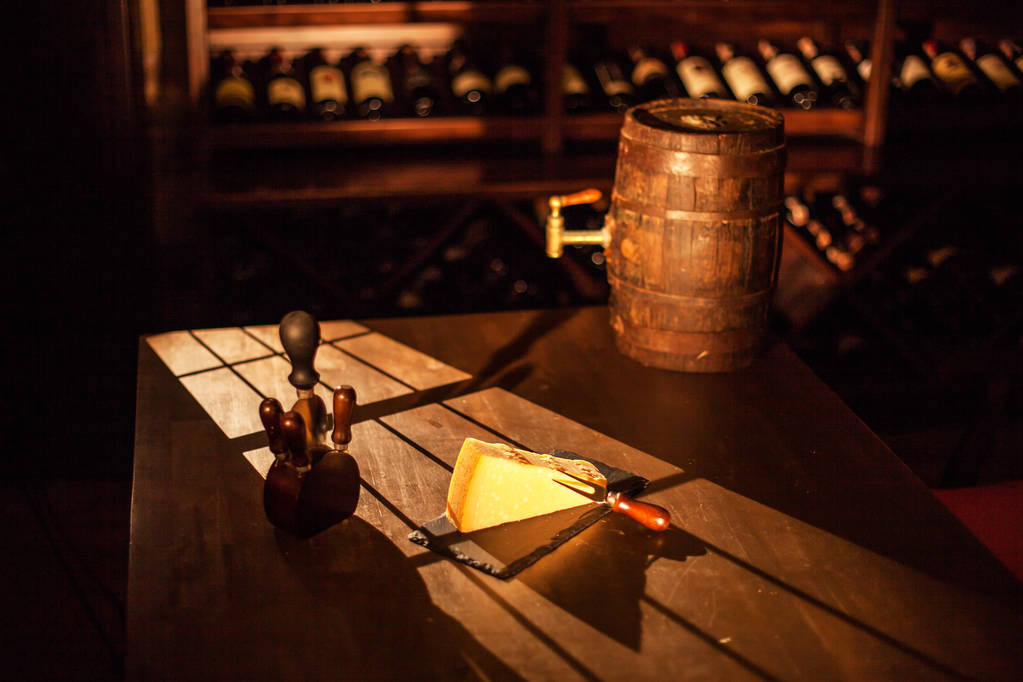 Composición si pieza de queso y accesorios colocados junto a la barrica de vino de madera en una mesa de madera en una bóveda de vino
. - Foto, imagen