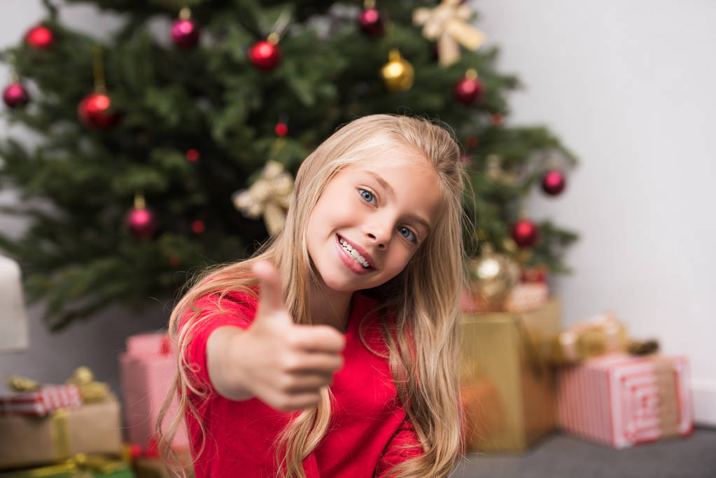kind met duim omhoog bij de kerstboom  - Foto, afbeelding