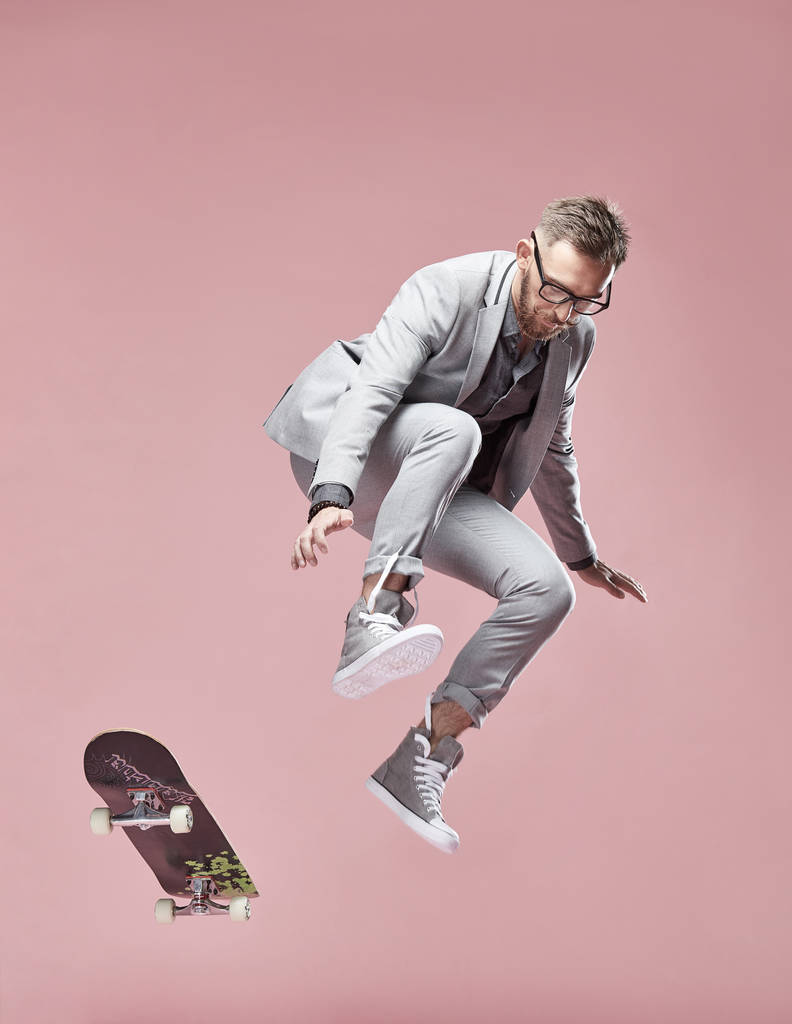Όμορφος σοβαρές νεαρός με γυαλιά, καστανά μαλλιά και γένια, φορώντας ελαφριά γκρι κοστούμι και πάνινα παπούτσια, άλμα με το skateboard σε ανοιχτό ροζ φόντο  - Φωτογραφία, εικόνα
