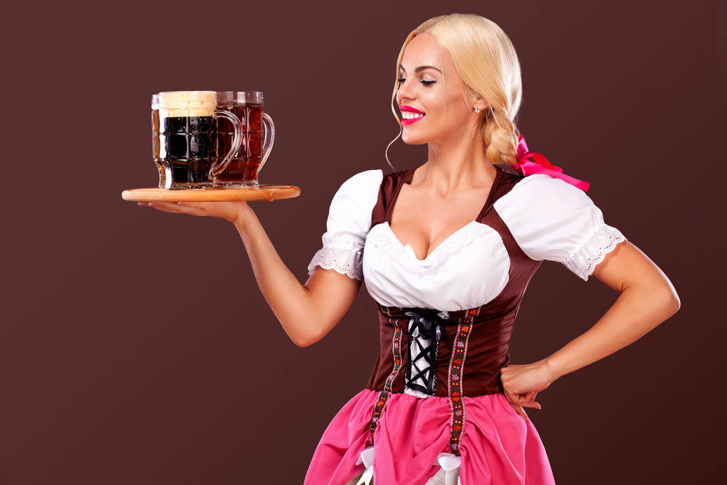 Портрет девушки с фестиваля Октоберфест - официантки в традиционном баварском платье, подающей большие кружки пива на коричневом фоне
. - Фото, изображение