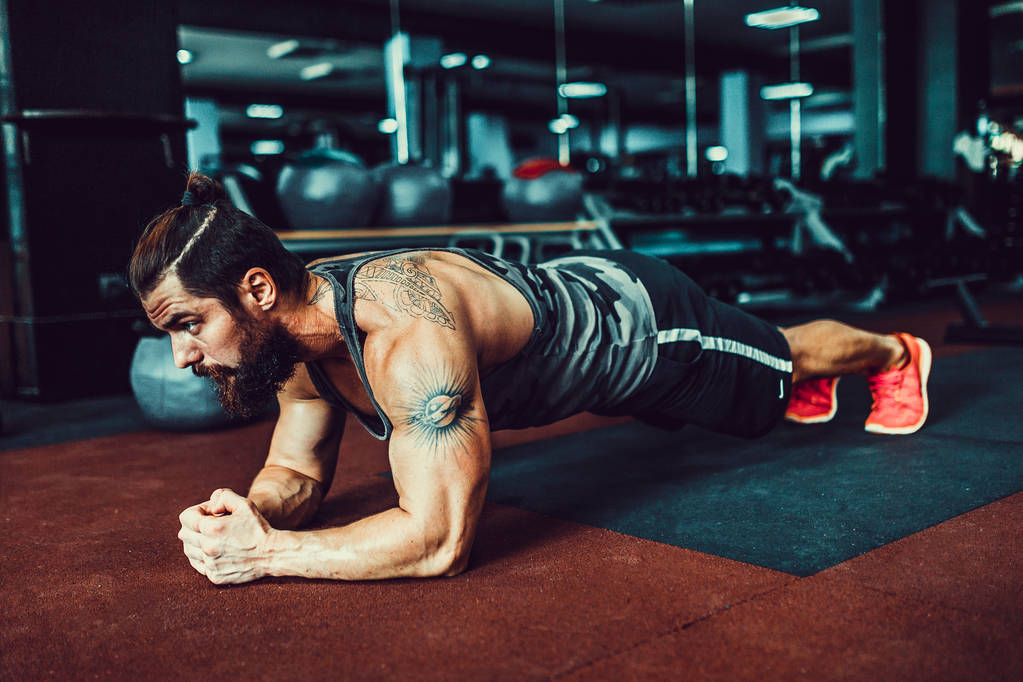 Muscoloso giovane uomo indossa abbigliamento sportivo e facendo posizione tavola durante l'esercizio sul pavimento in loft interno
 - Foto, immagini