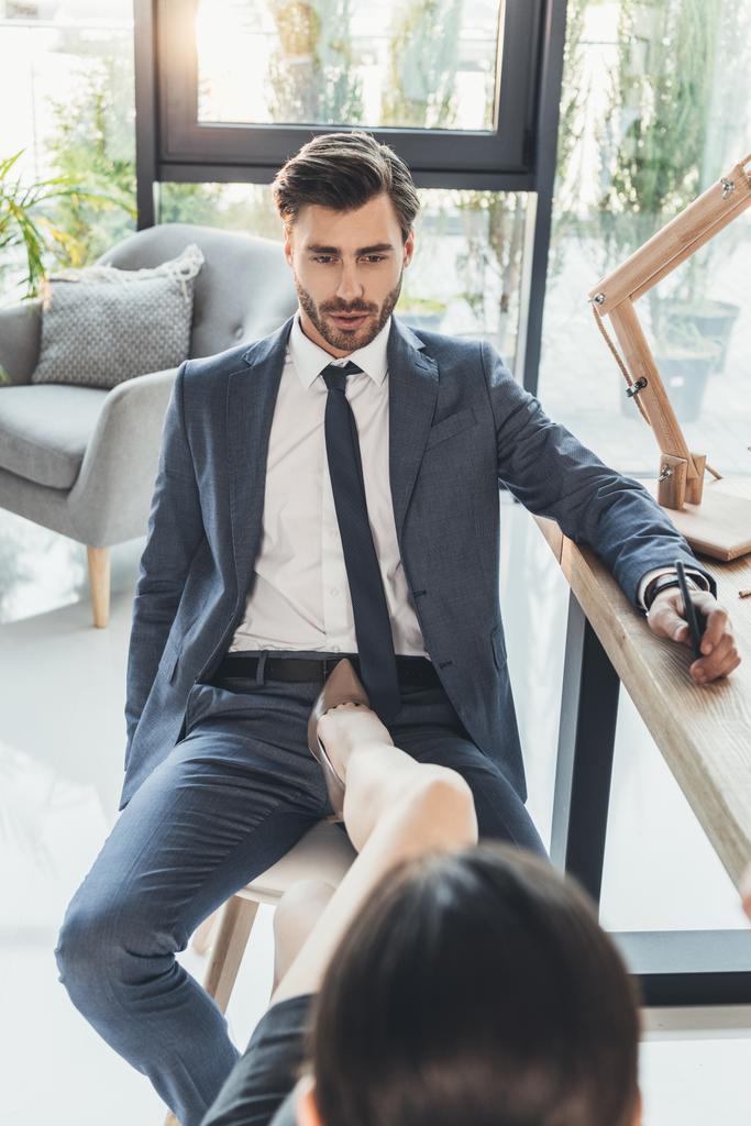 Жінка на високих підборах розміщує ногу на стільці між ногами молодого чоловіка в бізнесі
 - Фото, зображення