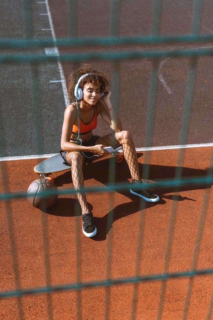 jonge Afro-Amerikaanse vrouw in sportkleding luisteren naar muziek in hoofdtelefoons en zittend op een longboard op sportveld - Foto, afbeelding