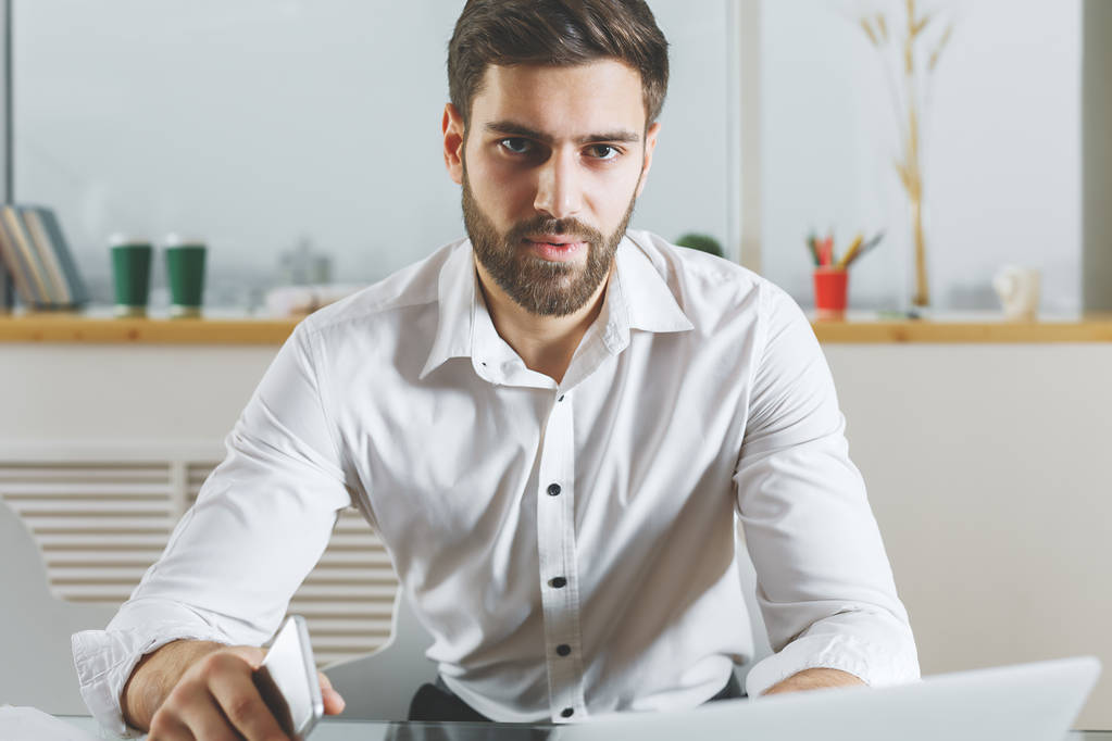 Porträt eines jungen europäischen Geschäftsmannes mit Smartphone in der Hand und Laptop am modernen Bürotisch. Arbeitskonzept  - Foto, Bild