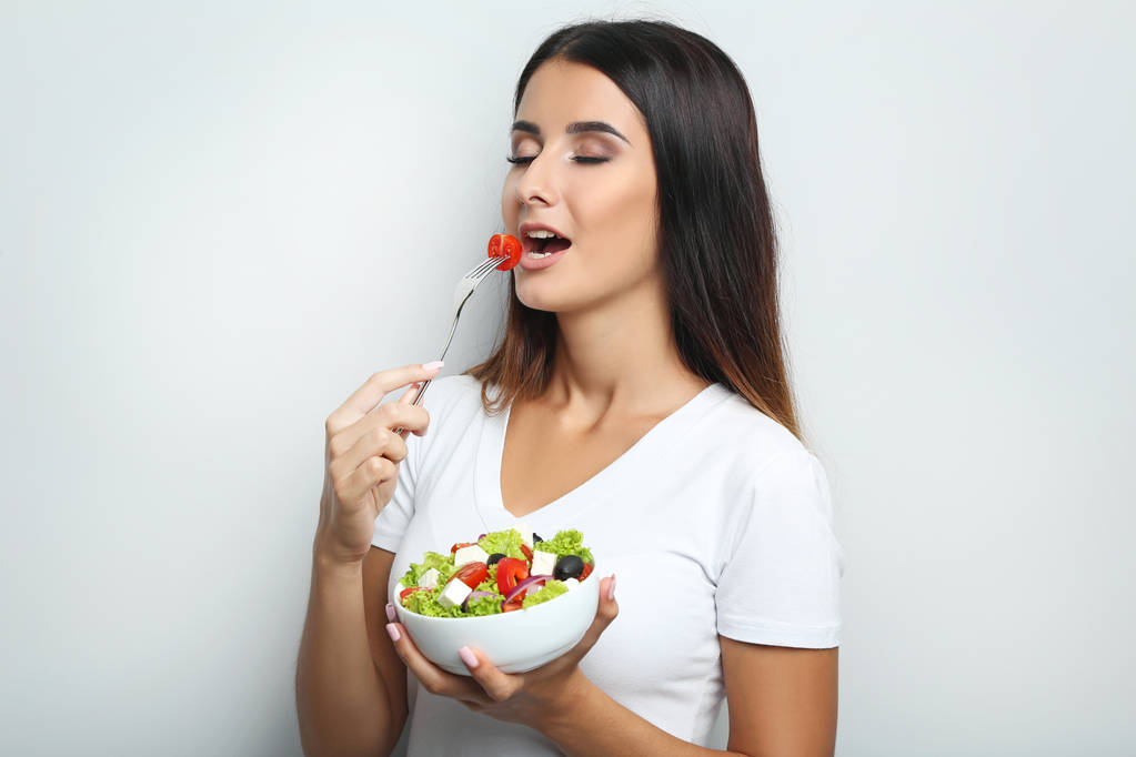 Jonge vrouw met plantaardige salade in kom op grijze achtergrond - Foto, afbeelding