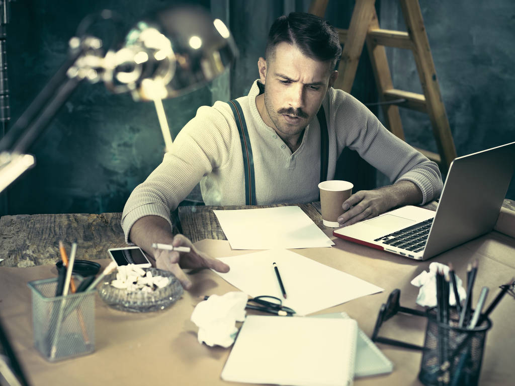 Красивый элегантный мужчина сидит за домашним столом, работает и использует ноутбук во время курения сигарет
 - Фото, изображение