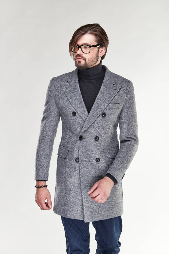 Attractive young sexy serious man model with beard and glasses posing in grey cashmere coat and dark blue pants in light studio  - Photo, Image