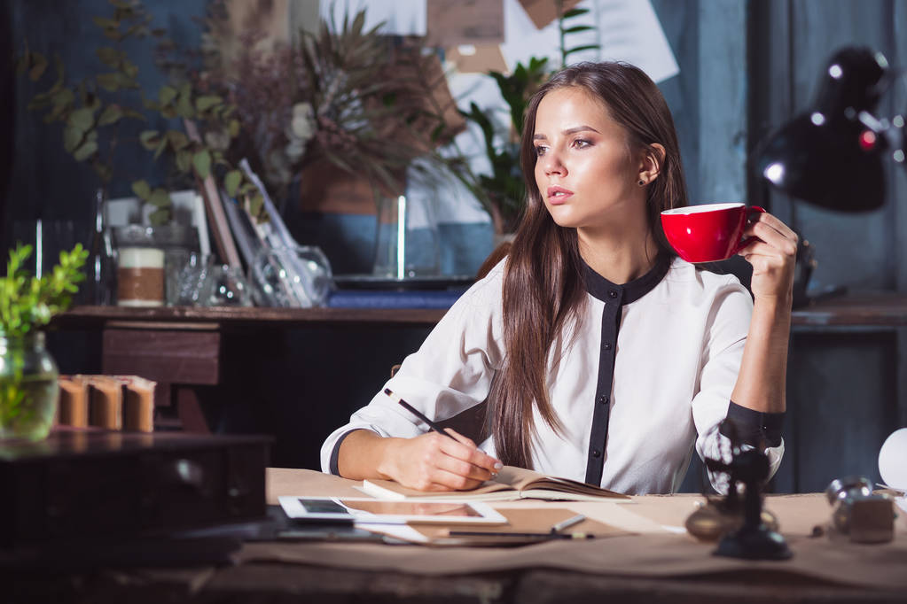 Νεαρή όμορφη γυναίκα που εργάζεται με φλιτζάνι καφέ και σημειωματάριο στο πατάρι γραφείο - Φωτογραφία, εικόνα