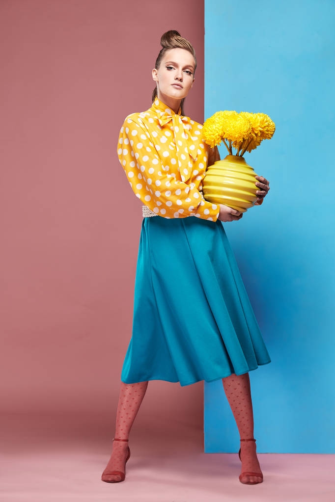Pretty young serious woman model wearing yellow blouse with white polka-dot, blue skirt and pink tights, holding yellow vase with yellow flowers and posing in studio with blue and pink background - Photo, Image