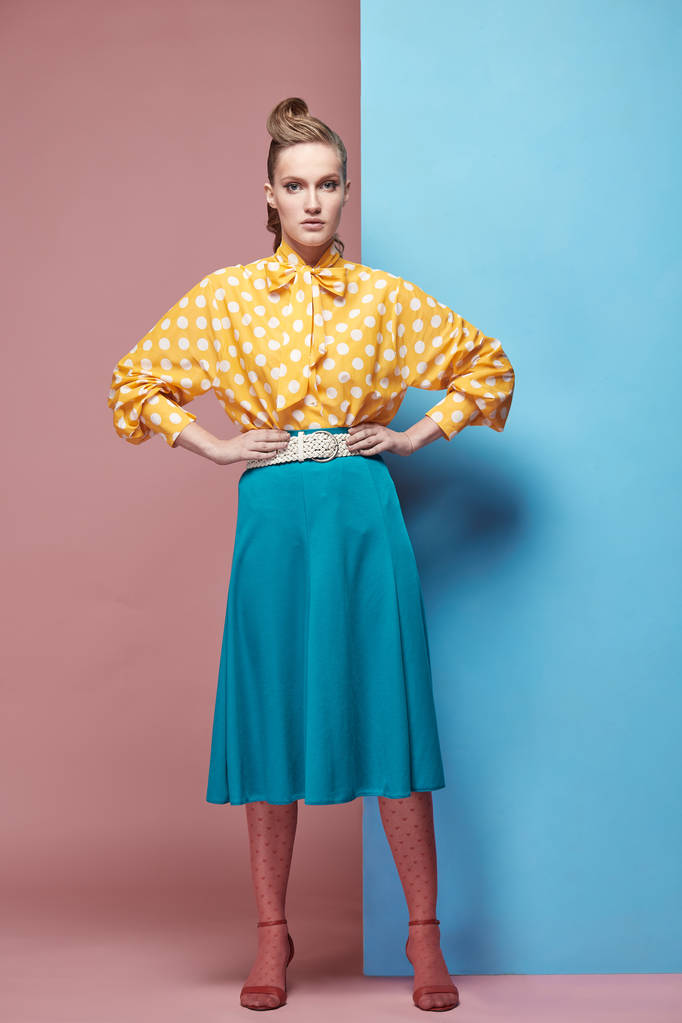 Bastante joven modelo de mujer seria con blusa amarilla con lunares blancos, falda azul y medias de color rosa y posando en el estudio con fondo azul y rosa
 - Foto, Imagen