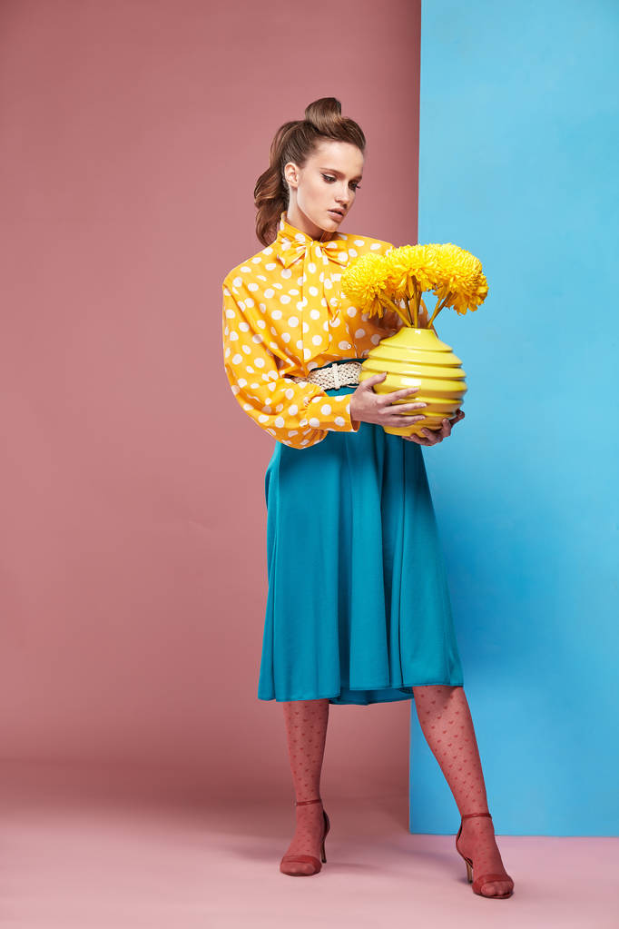 schöne junge ernsthafte Frau Modell trägt gelbe Bluse mit weißem Tupfen, blauem Rock und rosa Strumpfhose in Pin-up-Stil, hält Vase mit gelben Blumen und posiert im Studio mit blauem und rosa Hintergrund - Foto, Bild