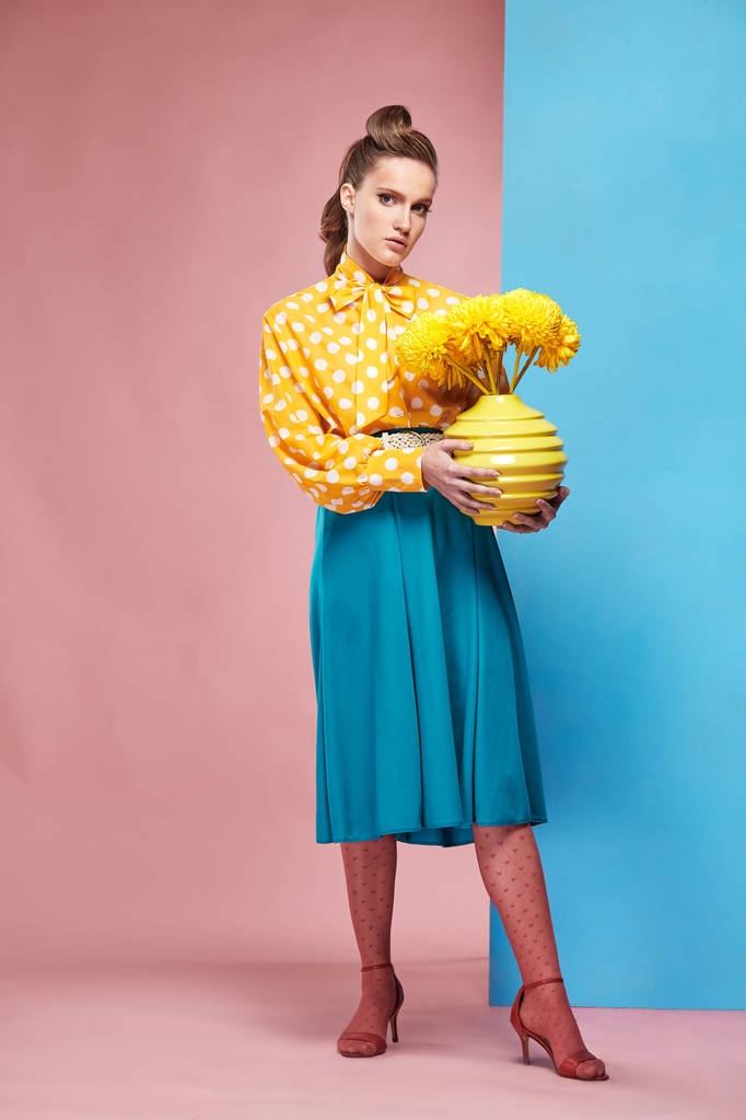 Красивая молодая сексуальная женщина модель в желтой блузке с белой горошек, синяя юбка и розовые колготки в стиле пин-ап, держа вазу с желтыми цветами и позируя в студии с синим и розовым фоном
 - Фото, изображение
