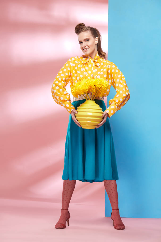 Incroyable jeune modèle de femme souriante sexy portant une blouse jaune à pois blancs, une jupe bleue et des collants roses en style pin-up, tenant un vase avec des fleurs jaunes et posant en studio avec un fond bleu et rose
  - Photo, image