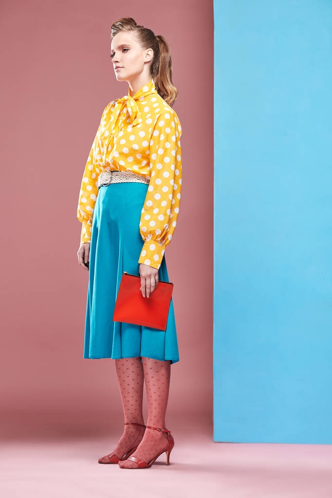 ピンナップ スタイルで、小さな赤いバッグを押し、青とピンクの背景のスタジオでポーズをとってホワイト ポルカ ドット、ブルーのスカートとピンクのタイツと黄色のブラウスを着てかなり若いセクシーな深刻な女性モデル  - 写真・画像