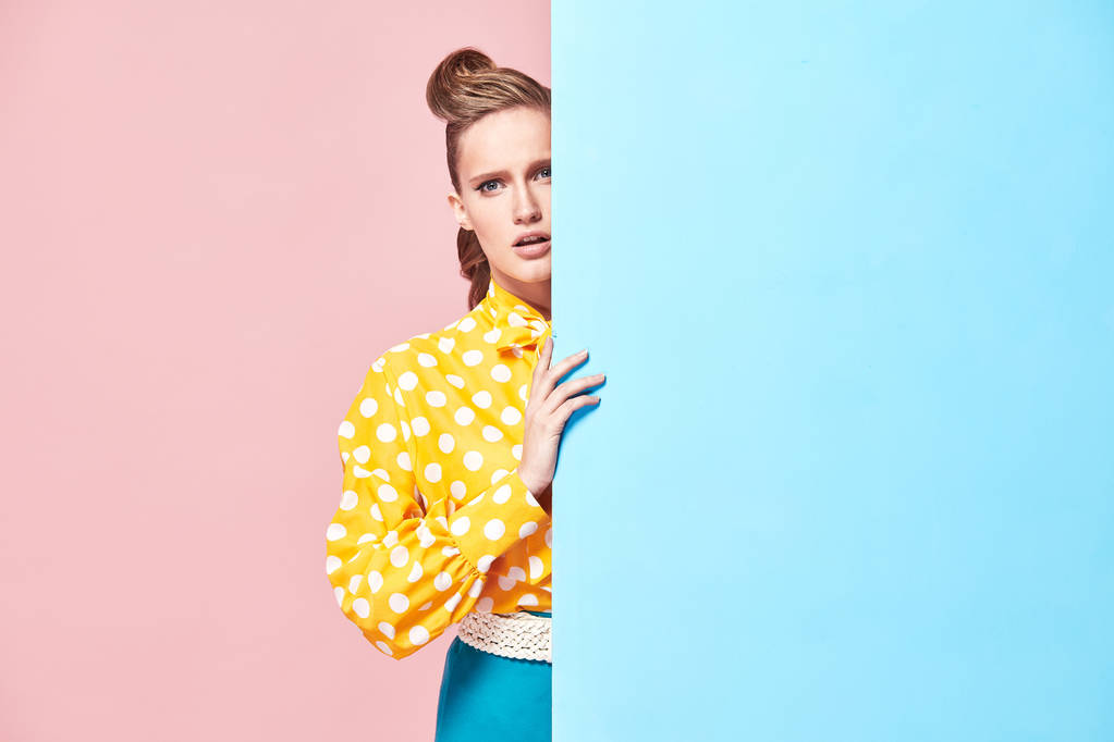 ピンナップ スタイルで、青色とピンク色の背景とスタジオで水色の壁の後ろに隠れている白い水玉模様水玉、青いスカートと黄色の魅力的な若い深刻な女性モデル着用の肖像画ブラウス  - 写真・画像