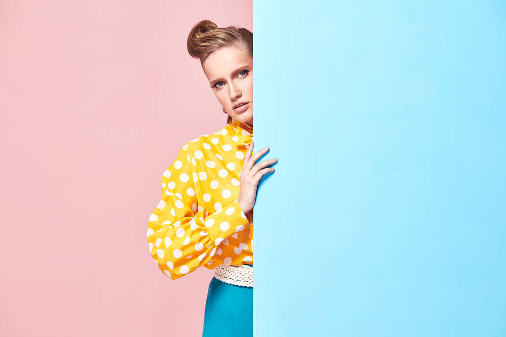 Ritratto di sorprendente giovane donna seria modello indossa camicetta gialla con pois bianco, gonna blu in stile pin-up, nascosto dietro parete azzurra in studio con sfondo blu e rosa
  - Foto, immagini