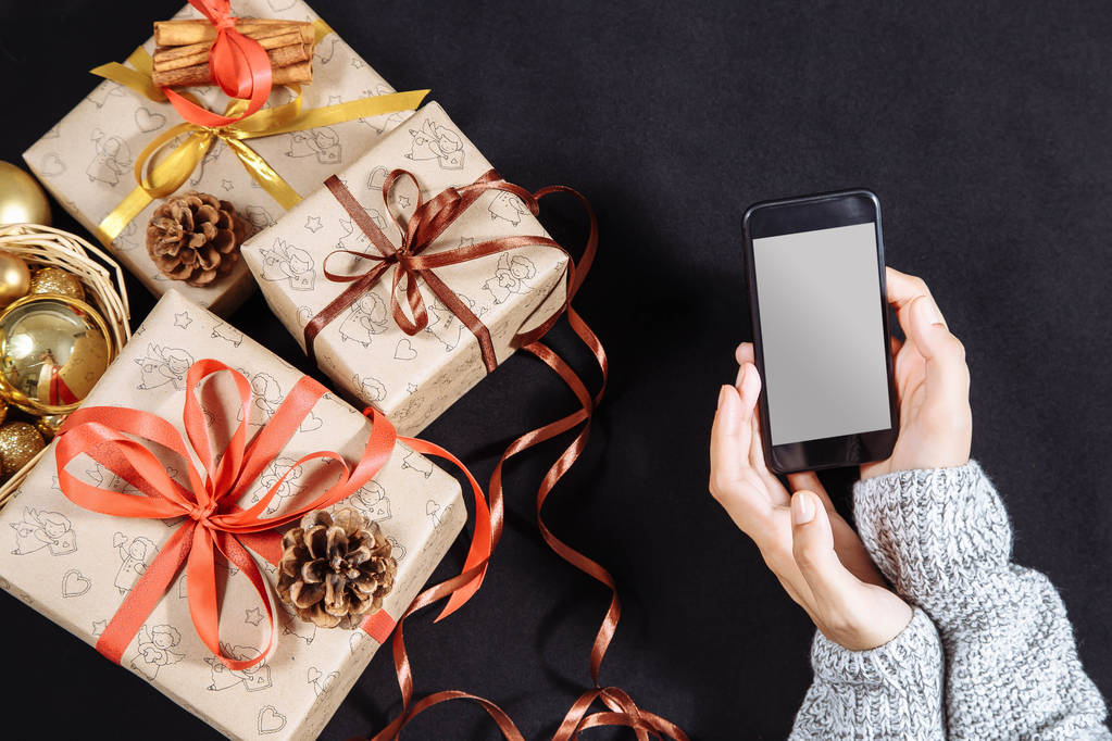 Manos femeninas sosteniendo el teléfono móvil sobre fondo oscuro con regalos de Navidad. Navidad y Feliz Año Nuevo composición. Piso tendido, vista superior
 - Foto, imagen
