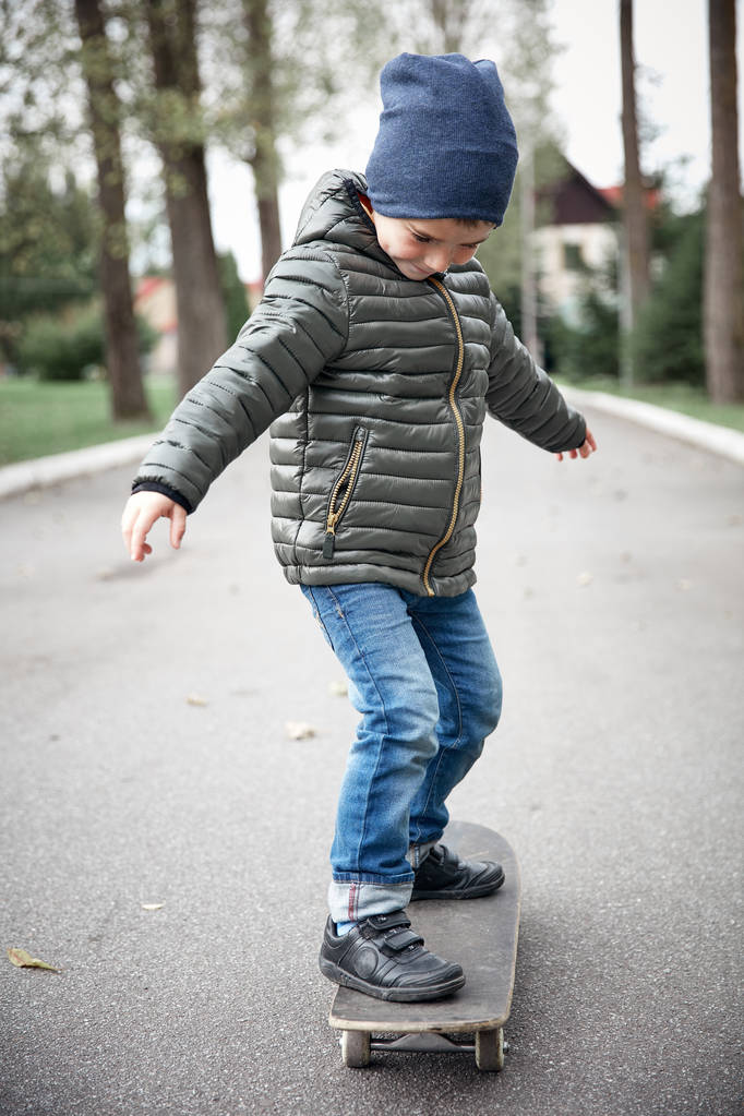 niedlich lustig lächelnder kleiner Junge in dunkelgrauer Jacke, dunkelblauer Mütze und blauer Jeans, der auf einem grauen Skateboard auf einem Asphaltweg im Freien fährt - Foto, Bild