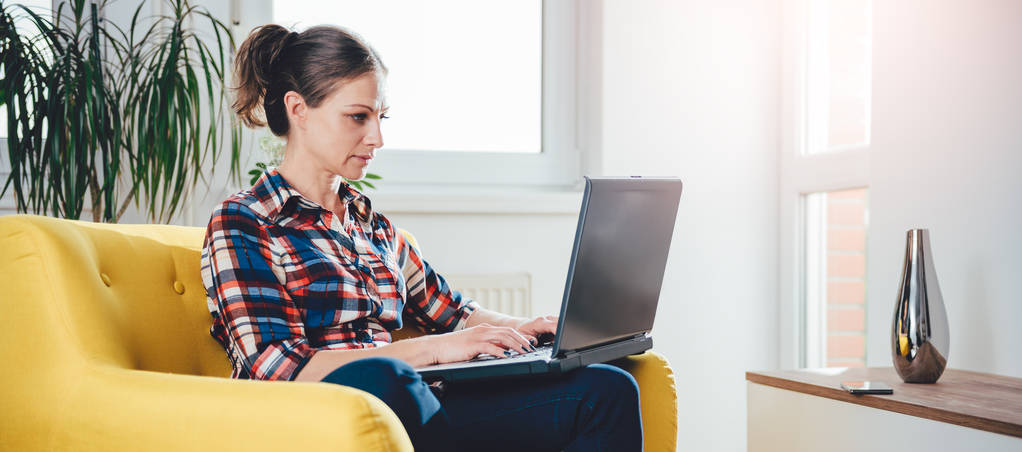 Жінка сидить на жовтому дивані у вітальні і використовує ноутбук
 - Фото, зображення