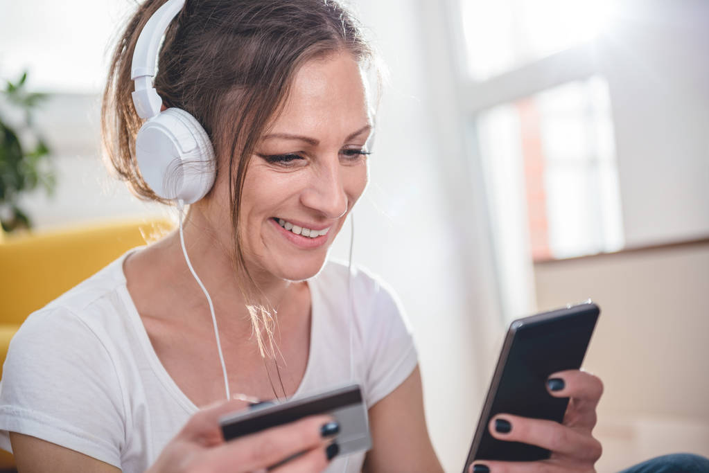 Nainen yllään valkoinen paita ja kuulokkeet ostoksia verkossa älypuhelimella ja luottokortilla
 - Valokuva, kuva