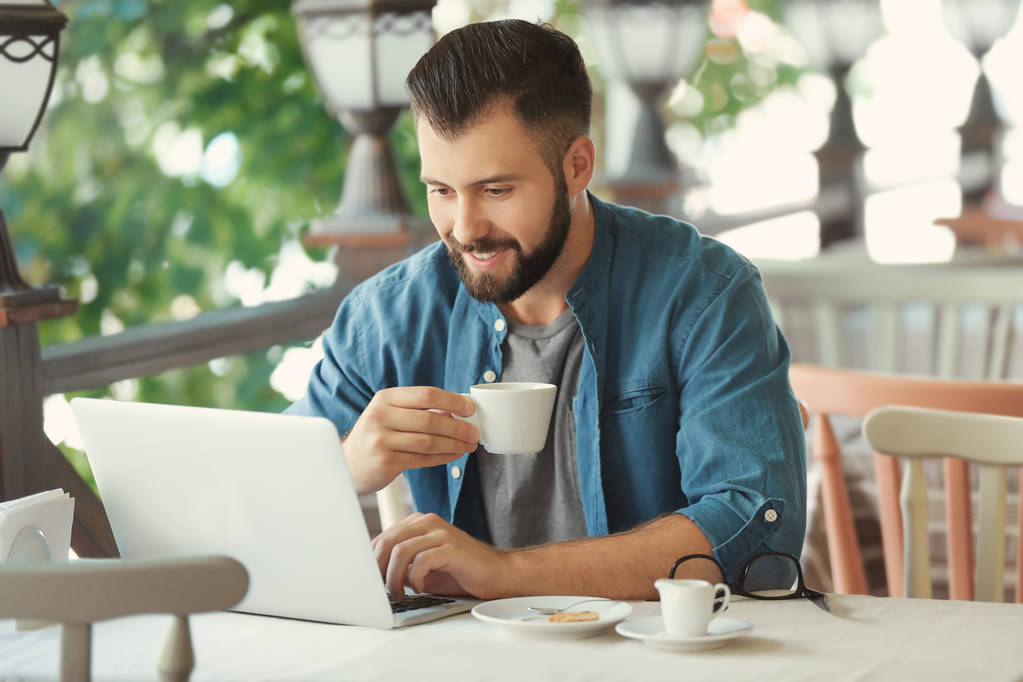 Молодой блоггер пьет кофе и пользуется ноутбуком в кафе на открытом воздухе
 - Фото, изображение