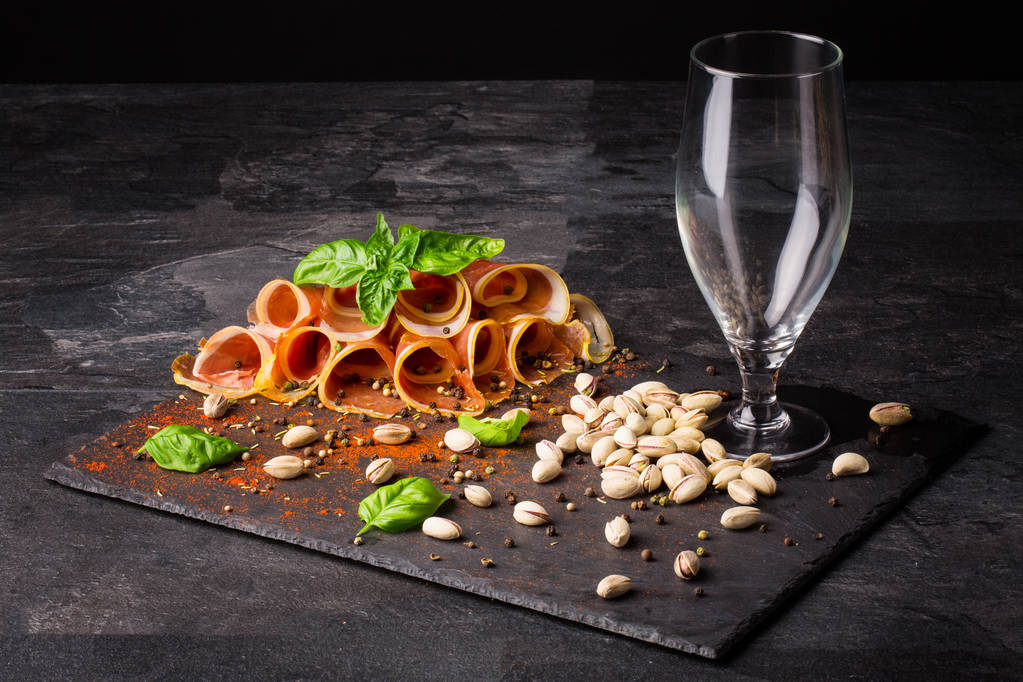 Close-up of an empty, tall and transparent glass and different tasty snacks on a dark brown cutting desk. Beautiful red ham, green basil leaves, salty pistachios and spices on a black background. - Photo, Image