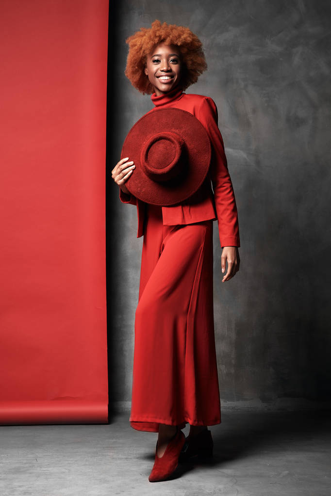 Молода красива чорна сексуальна усміхнена жінка в червоних штанях, піджак і взуття, тримає капелюх в руці, позує в студії з червоним і сірим фоном
 - Фото, зображення