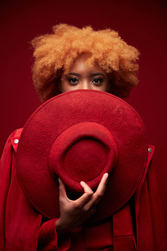Retrato de jovem muito preto sexy sério modelo mulher com cabelo curto encaracolado vermelho vestindo camisola vermelha, segurando chapéu vermelho e posando em estúdio no fundo vermelho
 - Foto, Imagem