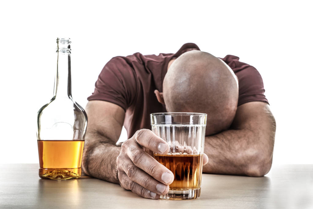 П'яний чоловік зі склянкою і пляшкою алкогольного напою сидить за столом на білому тлі
 - Фото, зображення