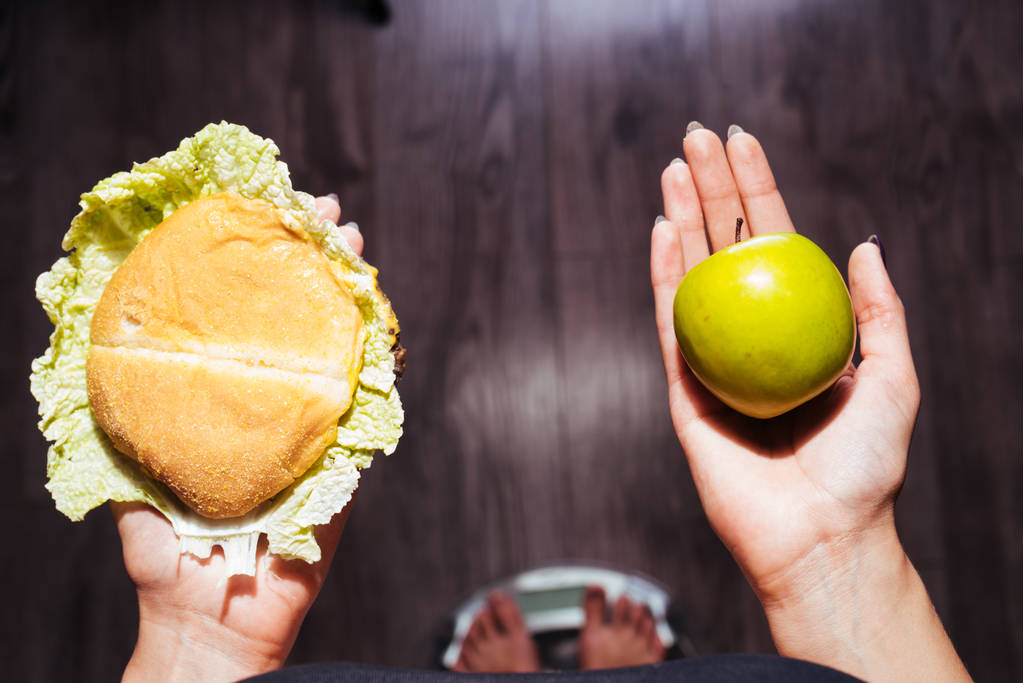 mladá dívka chce zhubnout, stojí na váze, má apple užitečné a škodlivé burger - Fotografie, Obrázek
