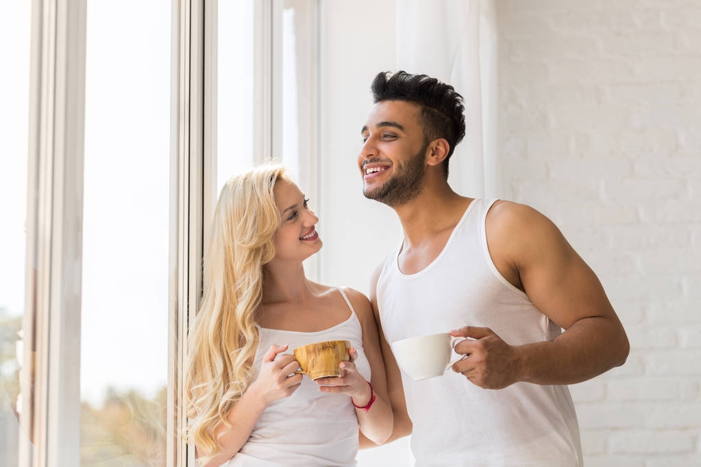 Młoda para piękny stojak w pobliżu duże okno, pić rano filiżanka kawy, szczęśliwy uśmiech Hiszpanie mężczyzna kobieta - Zdjęcie, obraz
