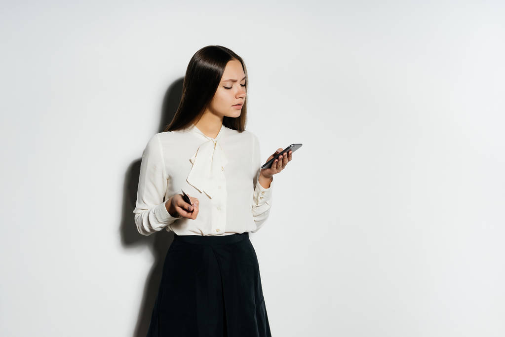 ein junges modernes ernstes Mädchen in weißem Hemd hält einen goldenen Bitcoin in der Hand, blickt in ihr Smartphone und studiert Kryptowährung - Foto, Bild
