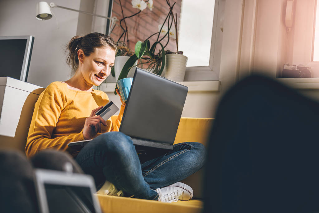 Жінка в жовтому светрі сидить на жовтому дивані вдома і купує онлайн з кредитною карткою
.  - Фото, зображення