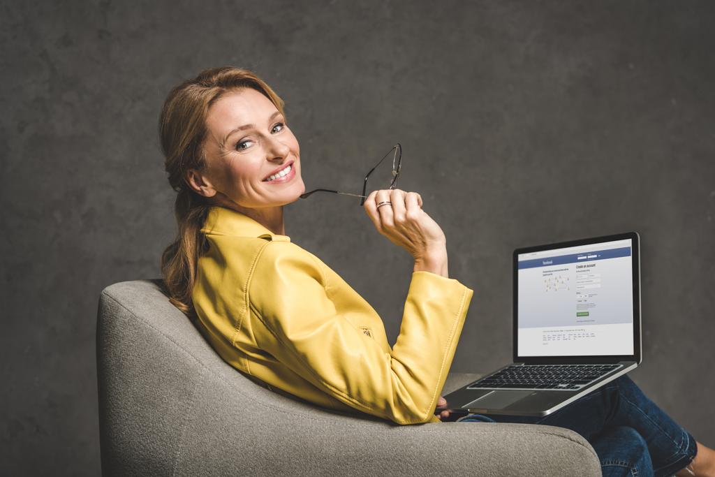 χαμογελώντας ώριμη γυναίκα χρησιμοποιώντας φορητό υπολογιστή με την ιστοσελίδα στο facebook και να κάθεται στην πολυθρόνα - Φωτογραφία, εικόνα