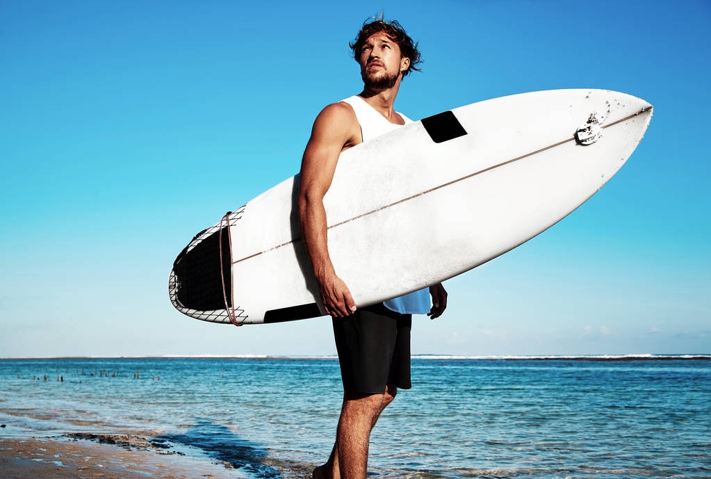 Портрет красивого хіпстера в сонячній моді модель чоловіка-серфера в повсякденному одязі, що йде з дошкою для серфінгу на синьому океані та фоні неба
 - Фото, зображення