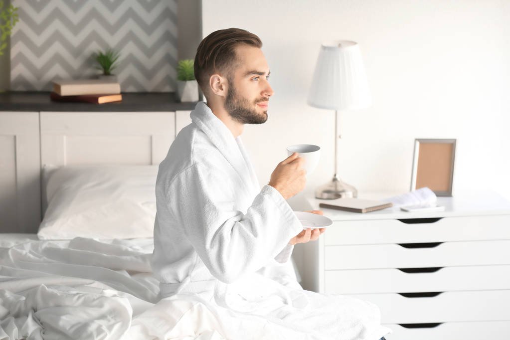自宅のベッドに座ってコーヒーを飲みながらハンサムな若い男の朝 - 写真・画像