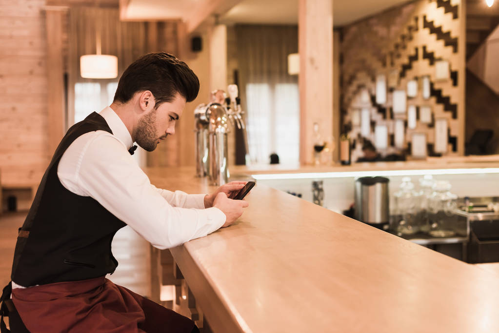 Офіціант сидить за барною стійкою зі смартфоном
 - Фото, зображення
