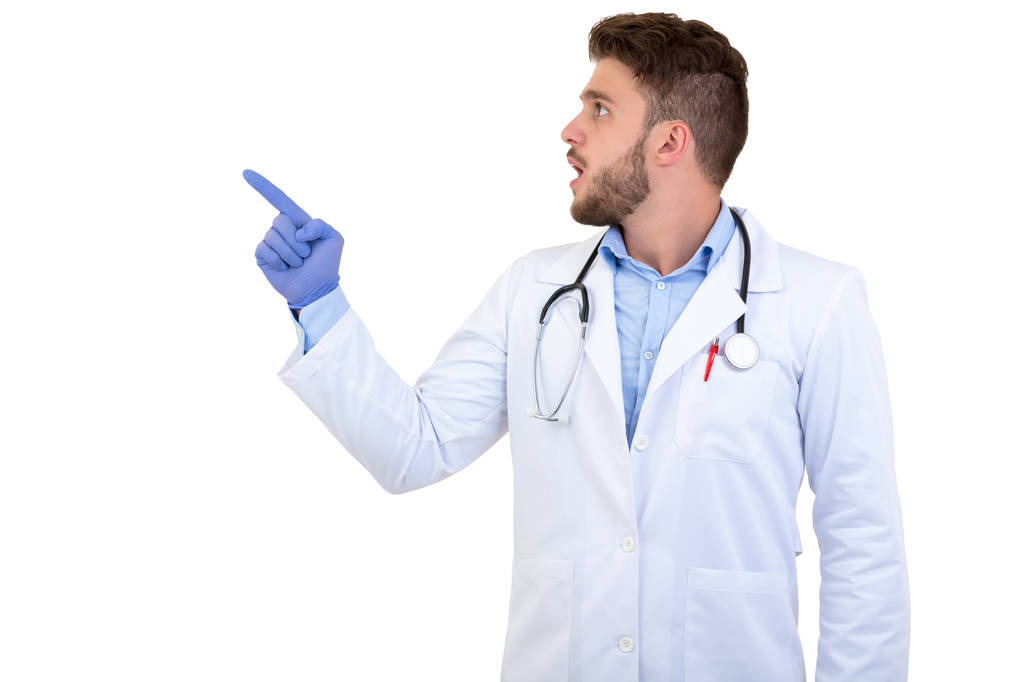 Porträt eines jungen lächelnden männlichen Arztes, der isoliert auf weißem Hintergrund mit dem Finger wegzeigt - Foto, Bild