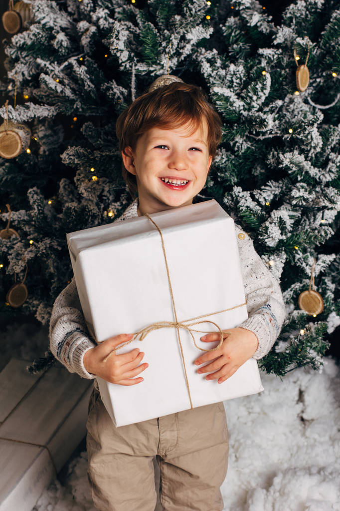Karácsonyi portré egy fiatal fiú hangulatos légkör, a karácsonyfa körül. Aranyos kisgyermek gazdaság fehér díszdobozban. Függőleges fénykép, másol cpace - Fotó, kép