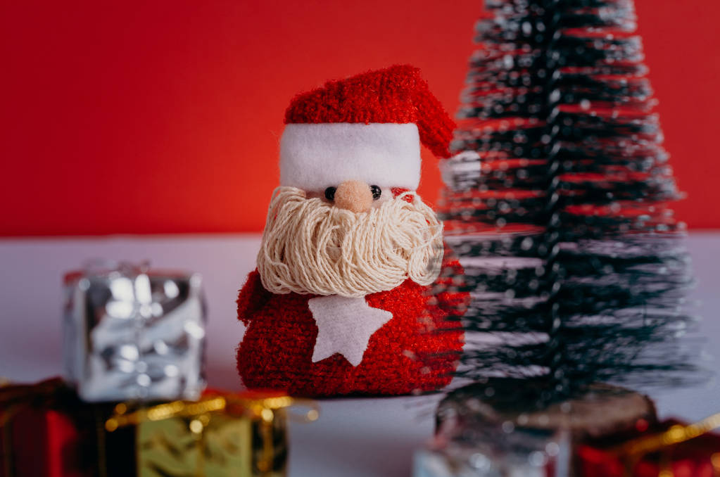 Милая игрушка Санта-Клауса спрятана между подарками и елкой, с красным фоном
. - Фото, изображение