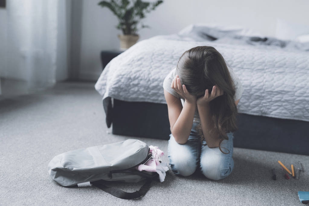 Чорноволоса дівчина сидить на підлозі поруч з її сірим рюкзаком і плаче над її обличчям руками
 - Фото, зображення