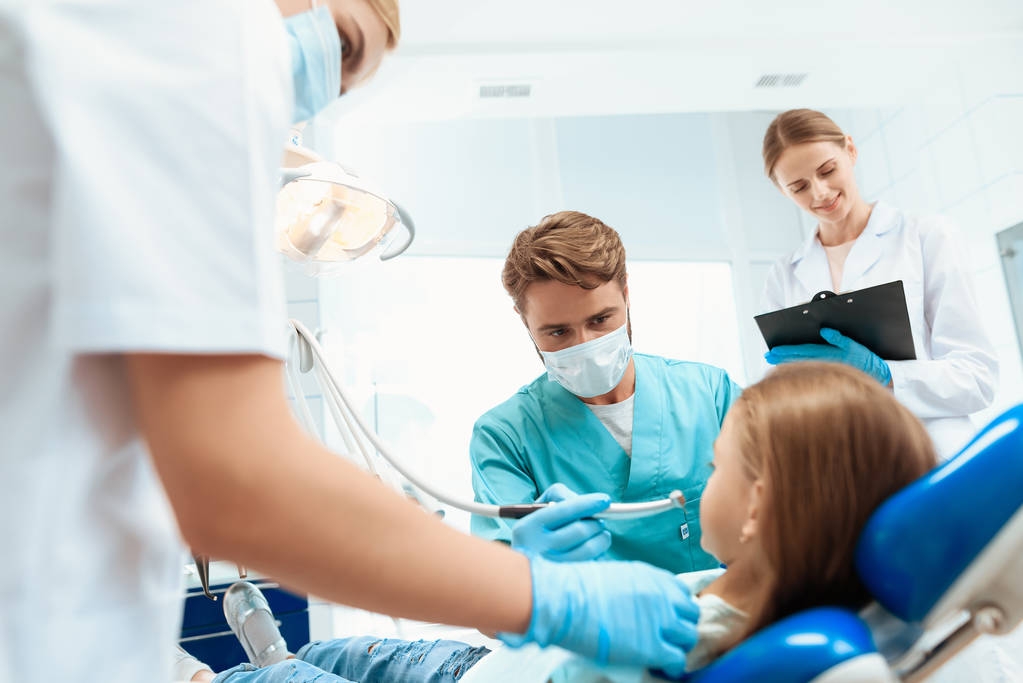Ένας οδοντίατρος ετοιμάζεται να τη θεραπεία των δοντιών ενός μικρού κοριτσιού. Υπάρχουν νοσηλευτές γύρω. Το κορίτσι που χαμογελά - Φωτογραφία, εικόνα