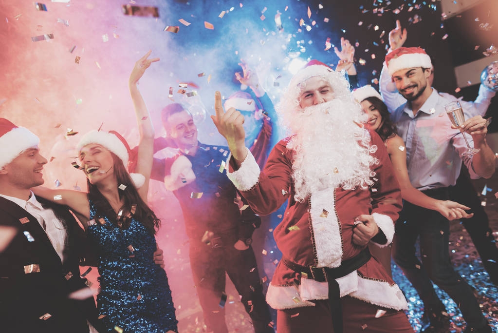 Um homem vestido de Papai Noel se diverte em uma festa de Ano Novo. Juntamente com ele ter amigos divertidos. Em torno deles voa confete, contra um fundo de névoa multi-colorida. As pessoas são muito alegres
. - Foto, Imagem
