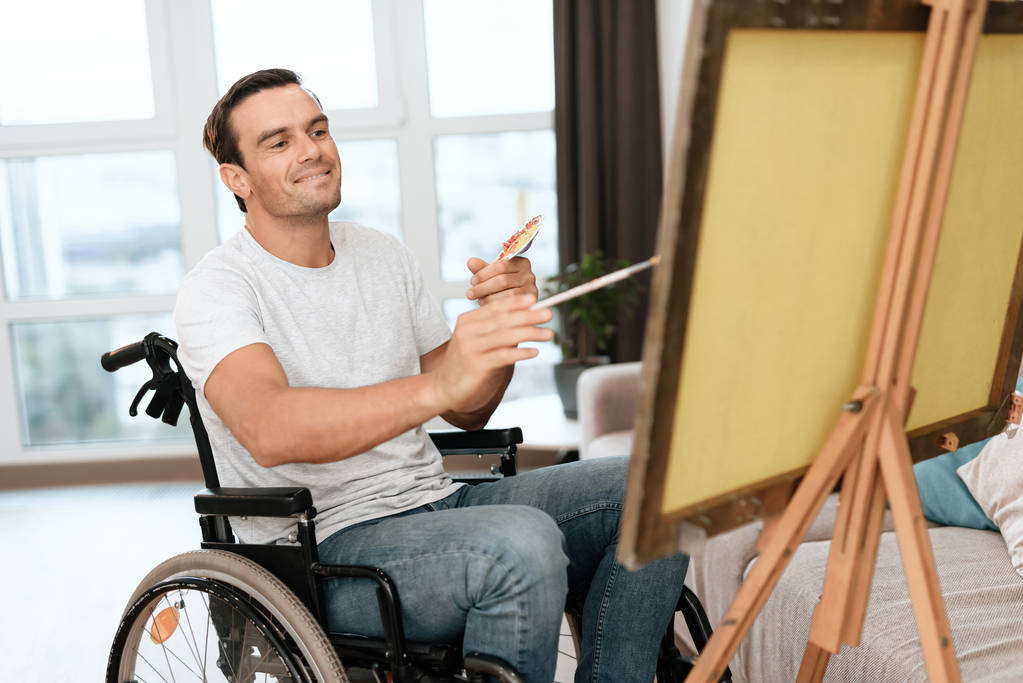 Το άτομο με αναπηρίες είναι κάθεται σε μια αναπηρική καρέκλα απέναντι από το καβαλέτο. Αντλεί μια εικόνα. Είναι στο μεγάλο φωτεινό σαλόνι του. Χαμογελάει. - Φωτογραφία, εικόνα