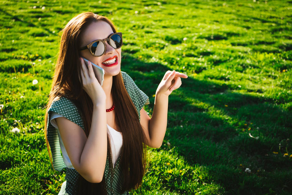 Ευτυχισμένη γυναίκα γυαλιά επισημαίνοντας κάτι στο γρασίδι, χρησιμοποιεί κινητά - Φωτογραφία, εικόνα
