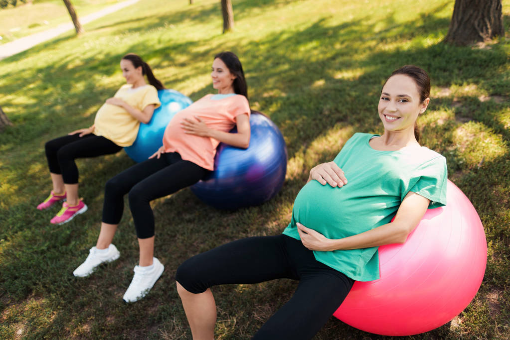 Três mulheres grávidas deitam-se em bolas de ioga no parque. Eles seguram suas barrigas
 - Foto, Imagem