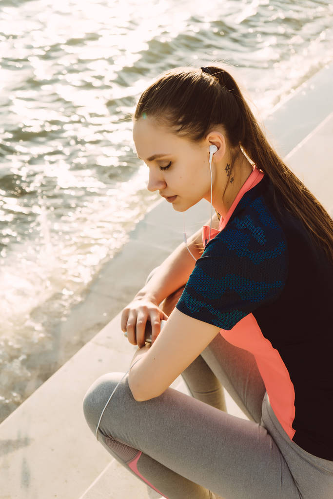 Dziewczyna usiadła na kroki na brzegu rzeki do słuchania muzyki w słuchawkach po treningu - Zdjęcie, obraz
