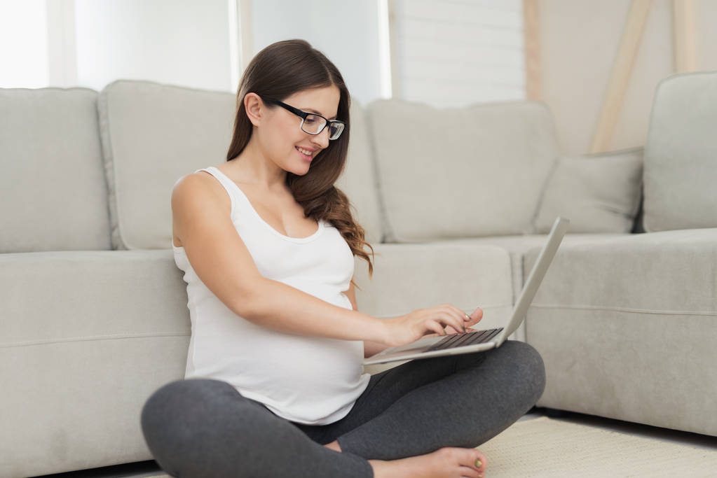 A pregnant woman sits on a light floor at home. She is sitting on the floor with her legs crossed and working on laptop - Photo, Image